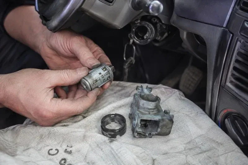 Hands holding a removed ignition cylinder beside a car steering column, with the housing and small parts on a cloth. — Ignition Lock Repair, Uxbridge